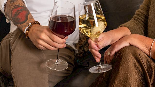 Two people clink wine glasses on a cozy couch, one wearing brown pants and boots, the other in a rust dress; relaxed, intimate cheers.