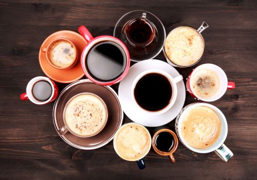 A top-down assortment of coffee drinks in colorful cups and mugs on a dark wooden table, featuring lattes, espresso, and creamers, arranged artfully.
