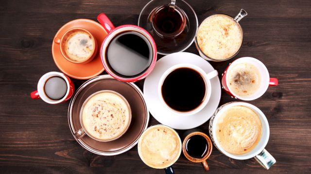 A top-down assortment of coffee drinks in colorful cups and mugs on a dark wooden table, featuring lattes, espresso, and creamers, arranged artfully.