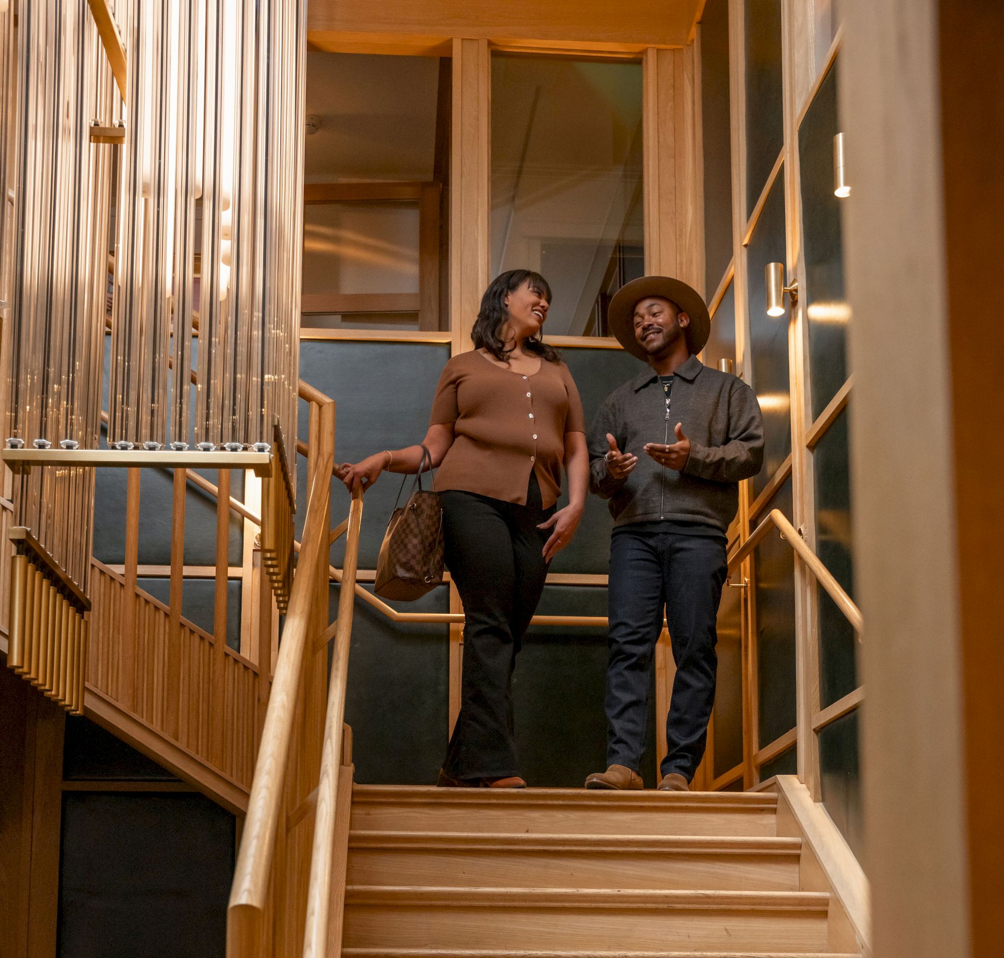 Two people stand on a wooden staircase landing, chatting as they look toward the camera, in a warm, modern interior.