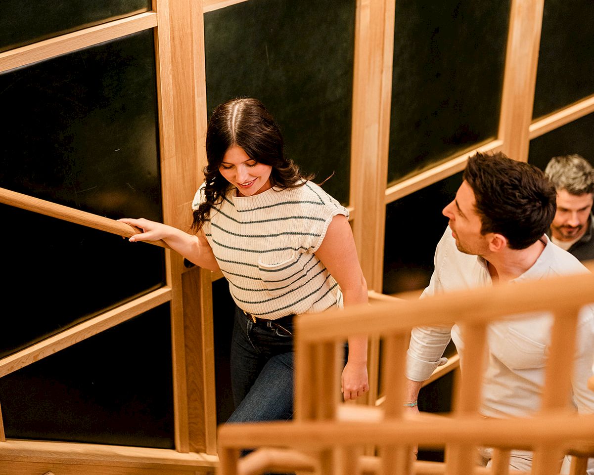 Three people ascend a wooden staircase by glass walls, with a woman in a striped shirt leading the way and two men following.