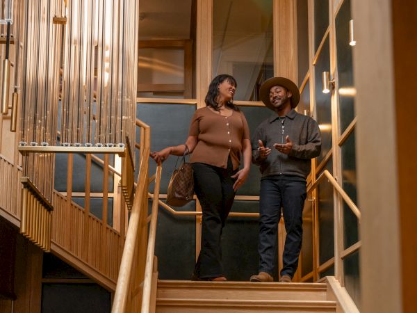 Two people stand on a wooden staircase indoors, chatting casually; the man wears a hat and denim, the woman shoulders bare with long dark hair.