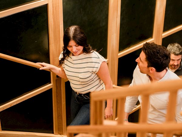 A woman leads a man up a wooden staircase, others following, inside a warmly lit, modern interior with large windows.