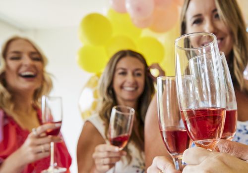 A group of friends toasting with pink drinks at a party, balloons in the background, smiling and celebrating together.