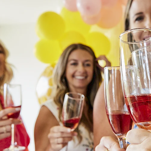 A group of friends toasting with pink drinks at a party, balloons in the background, smiling and celebrating together.