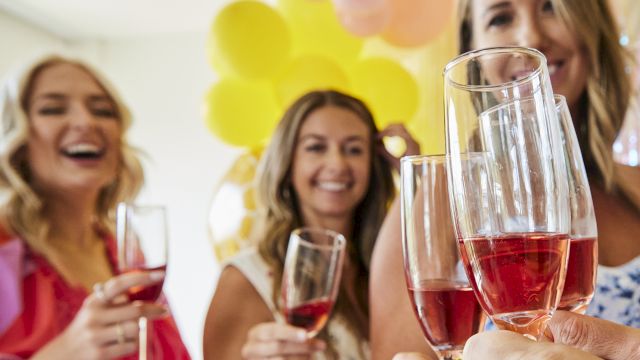 A group of friends toasting with pink drinks at a party, balloons in the background, smiling and celebrating together.