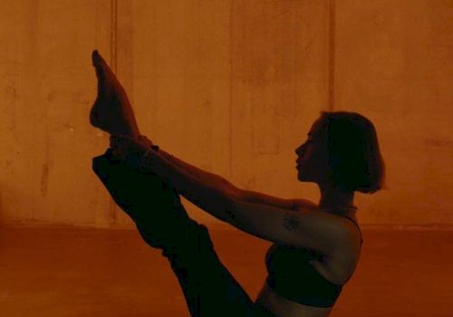 A person performing a boat pose in a dim, orange-lit room, balancing on their sit bones with legs lifted and arms reaching toward the feet.