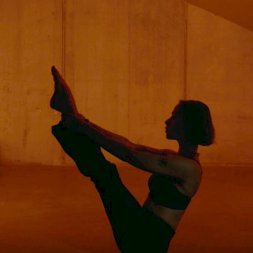 A person performing a boat pose in a dim, orange-lit room, balancing on their sit bones with legs lifted and arms reaching toward the feet.
