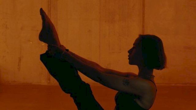 A person performing a boat pose in a dim, orange-lit room, balancing on their sit bones with legs lifted and arms reaching toward the feet.