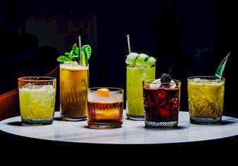 A row of colorful cocktails on a white round table, each glass with unique garnishes like mint, lime, and berries, set in a dim bar atmosphere.