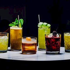 A row of colorful cocktails on a white round table, each glass with unique garnishes like mint, lime, and berries, set in a dim bar atmosphere.