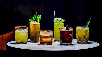 A row of colorful cocktails on a white round table, each glass with unique garnishes like mint, lime, and berries, set in a dim bar atmosphere.