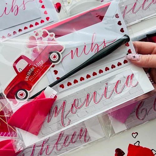 Valentine-themed gift set with pink cards, heart stickers, a small red toy car, and pink ribbon, all in clear packaging with names.