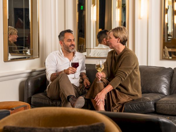 Two men sit on a couch in a stylish lounge, sipping wine and chatting warmly in a cozy, mirrored room.