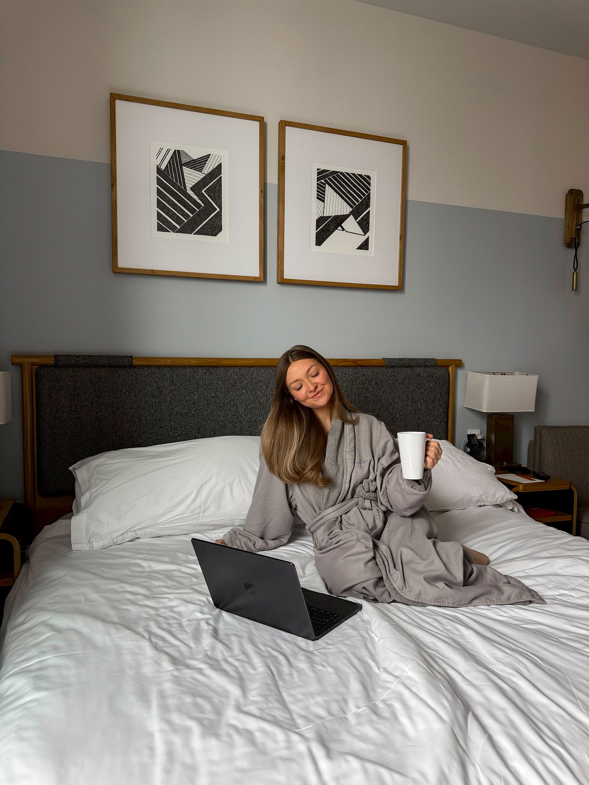 A woman sits on a bed with a laptop and coffee, wearing a gray robe, in a cozy hotel-like room with framed art above the headboard.