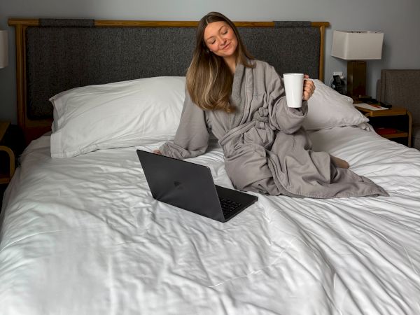 A woman sits on a bed with a laptop, holding a coffee cup, wearing a gray robe, in a cozy hotel room with framed abstract art above the headboard.