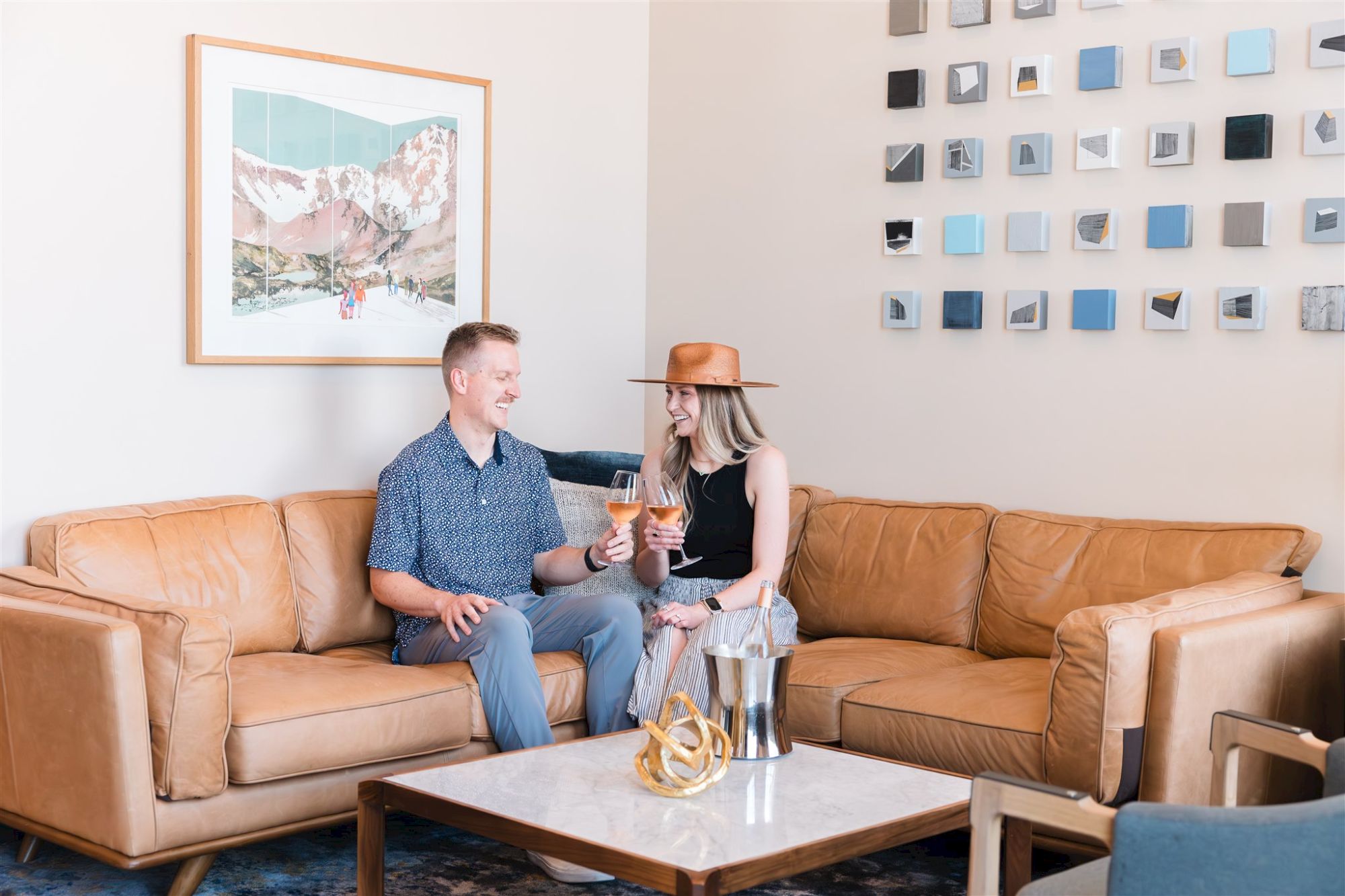 Two people sit on a tan leather L-shaped couch in a bright living room, talking and smiling as they share a drink; decor includes a wall grid and art.