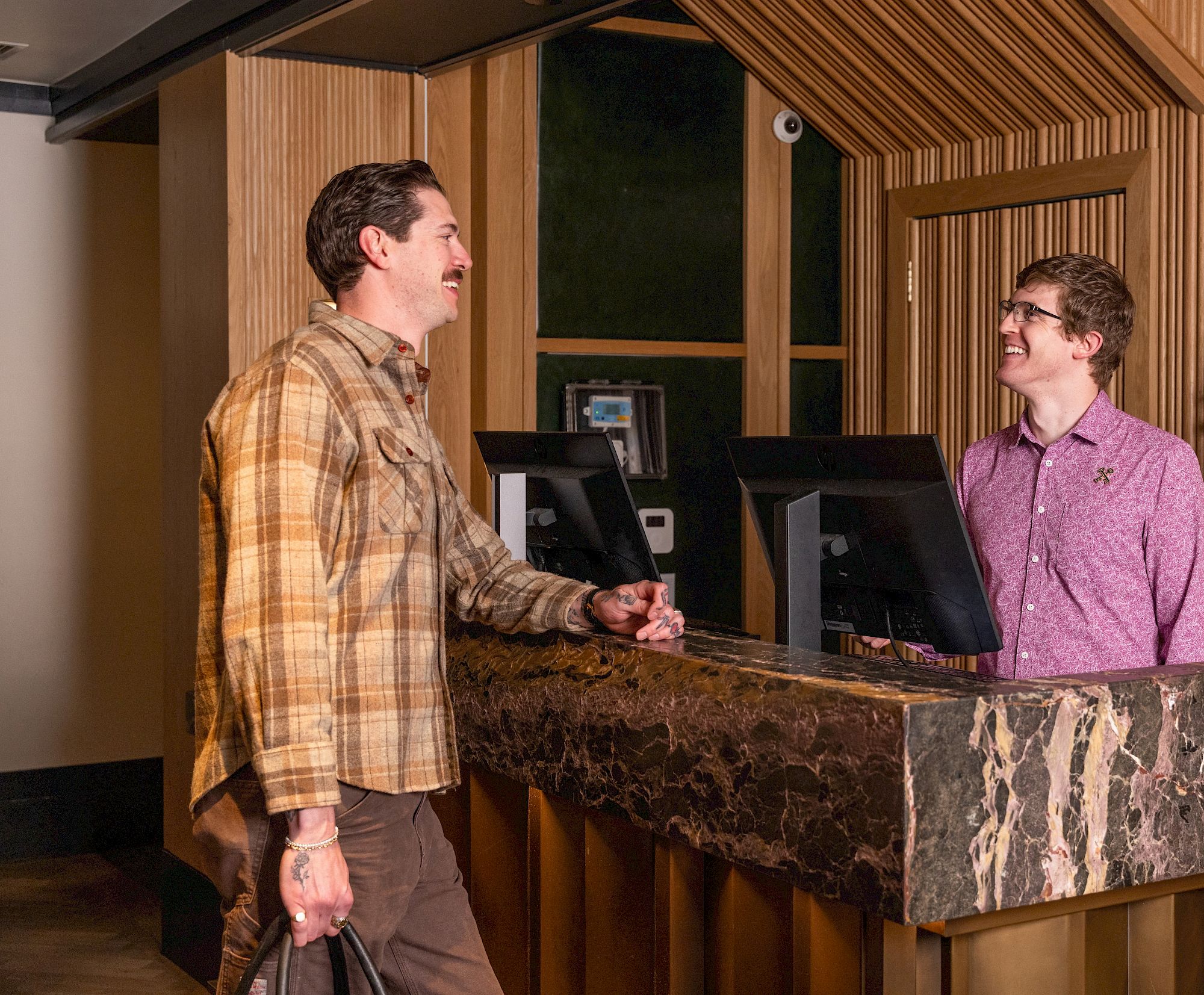 A man in a plaid shirt checks in at a hotel front desk with a smiling clerk on the marble counter.