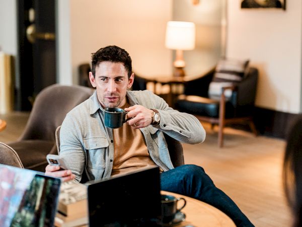 A man sits in a cozy living room, coffee mug in hand, working on a laptop at a round table with another laptop and phone nearby.