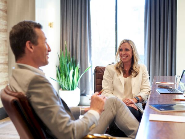 Two business colleagues chat in a bright office: a man in a suit sits relaxed, a woman in white blazer smiles across the desk as plants and laptops appear nearby.