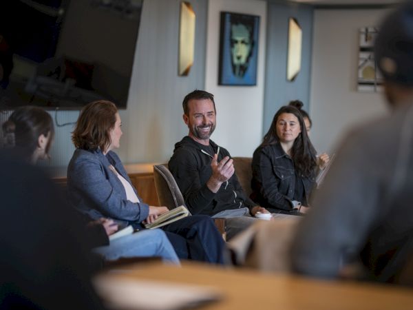 A group of people sit in a circle in a studio or meeting room, one man speaking while others listen attentively.