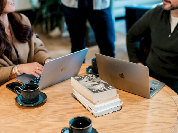 Two people sit at a round table with laptops, coffee cups, and a stack of books between them, collaborating in a casual office setting.