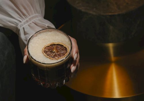 A person holds a dark beer with a dried citrus slice garnish, while a warm, brass lamp glows in the background, artfully lit.
