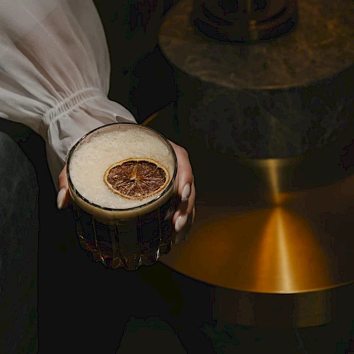A person holds a dark beer with a dried citrus slice garnish, while a warm, brass lamp glows in the background, artfully lit.