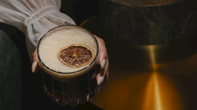 A person holds a dark beer with a dried citrus slice garnish, while a warm, brass lamp glows in the background, artfully lit.