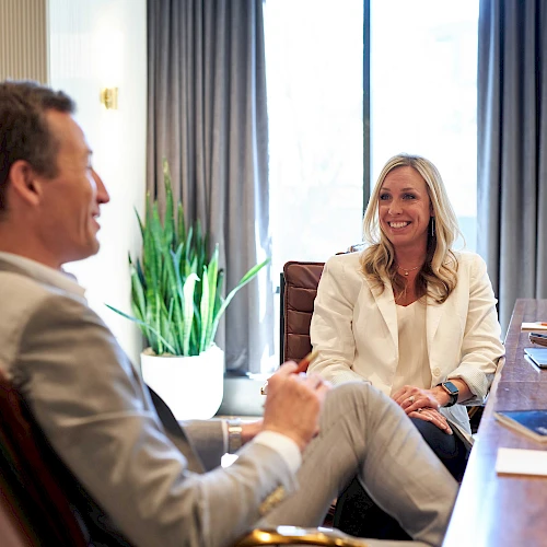 Two professionals in a bright office playfully chat during a meeting, smiling and seated across a conference table with laptops and papers.