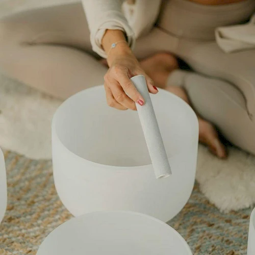 A person in beige lounge wear sits cross-legged, placing a rolled white stick into a white ceramic bowl on a textured rug, calm vibe.