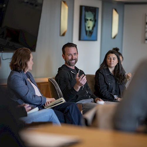 A group of people in a meeting room, one man speaking animatedly while others listen; a casual, collaborative discussion scene.