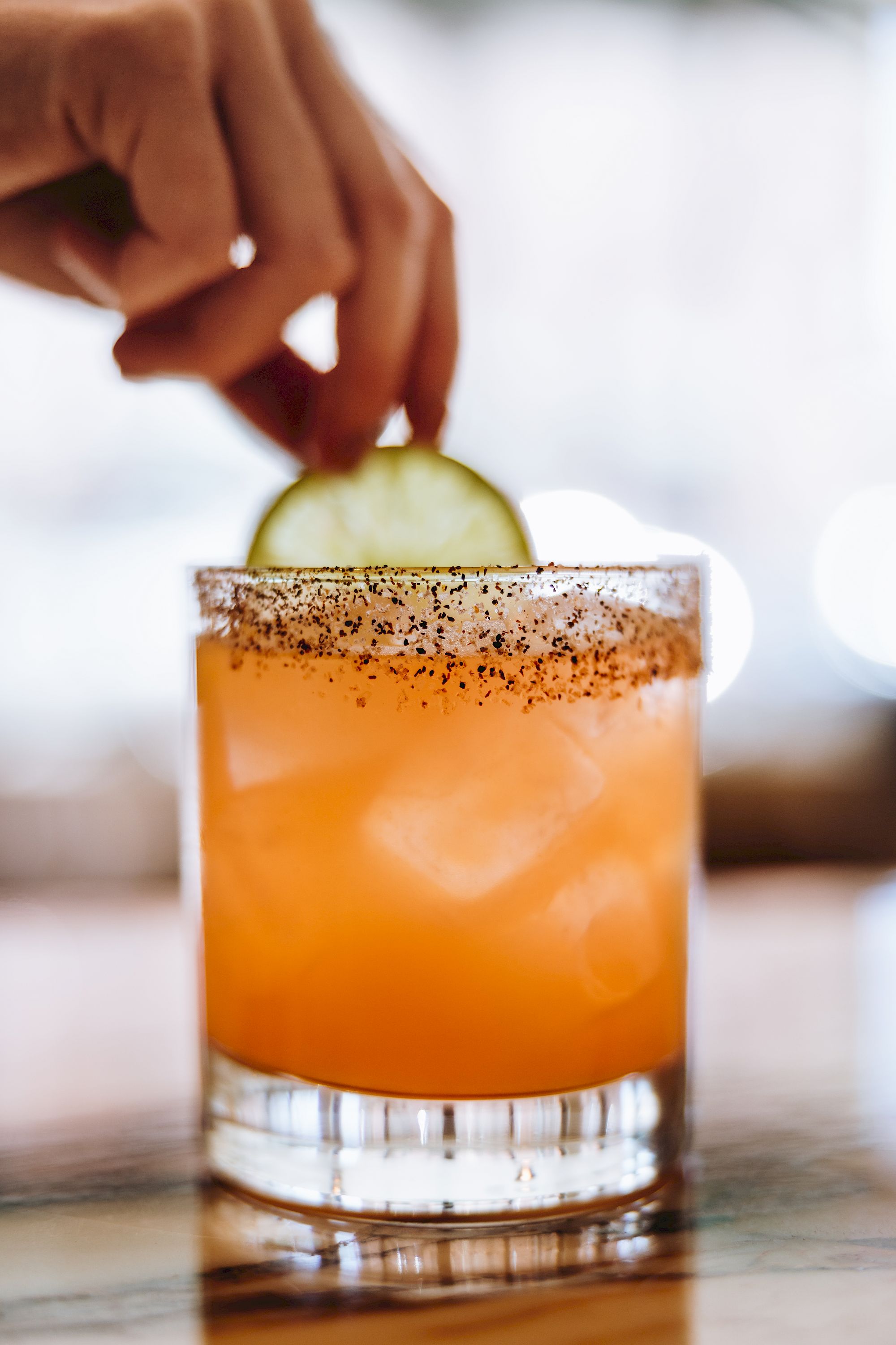 A hand adds a lime wheel to a frosty, orange cocktail with a salted rim, on a blurred bar backdrop, in a stylish, refreshing drink.