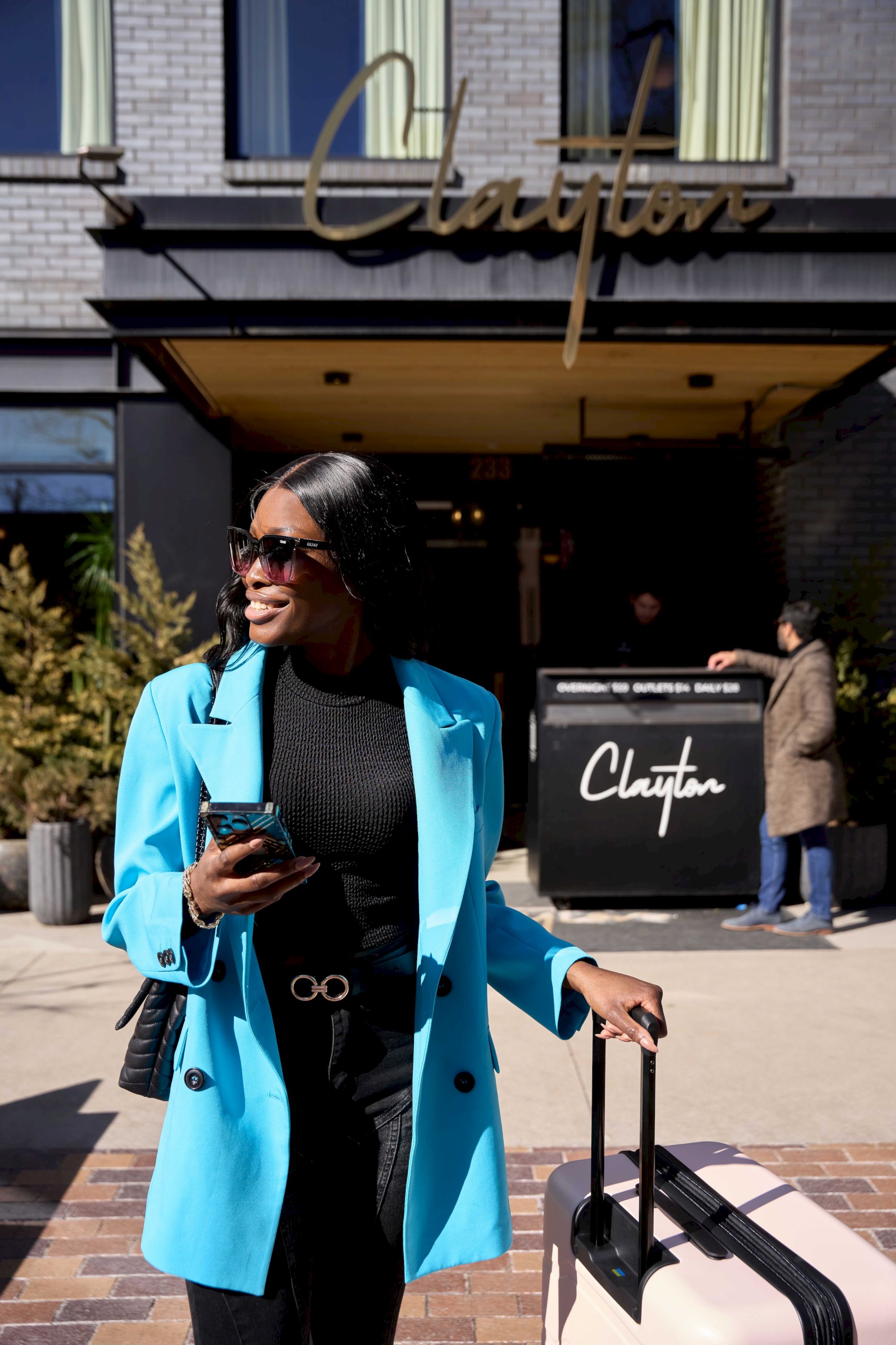 A stylish woman in a bright blue coat with sunglasses and a rolling suitcase stands outside a chic cafe, phone in hand, ready to travel.