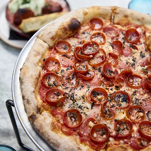 A pepperoni-tomato pizza with a golden crust, served on a metal tray with a side of dip and toppings nearby.