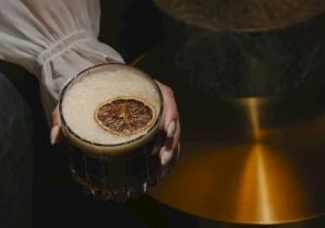 A person holds a cocktail glass with a dried orange slice garnish, in a moody, warm-lit setting.