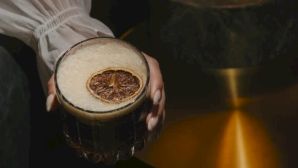 A person holds a cocktail glass with a dried orange slice garnish, in a moody, warm-lit setting.