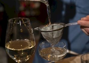 A glass of white wine and a person pouring through a fine mesh strainer into a smaller glass, with another glass nearby.