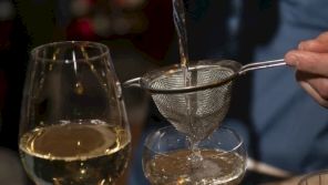 A glass of white wine and a person pouring through a fine mesh strainer into a smaller glass, with another glass nearby.