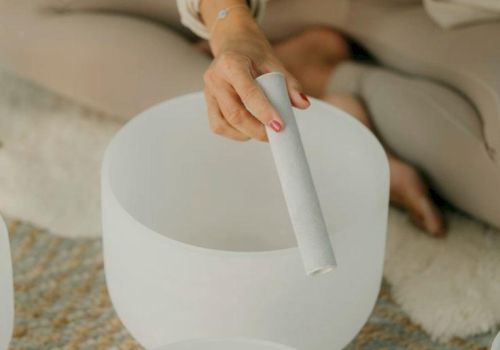 A person sits cross-legged, playing a white singing bowl instrument with a mallet, in a calm, minimalist setting.