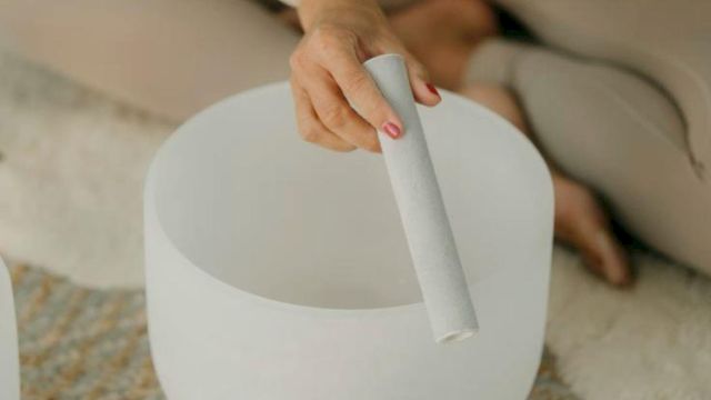 A person sits cross-legged, playing a white singing bowl instrument with a mallet, in a calm, minimalist setting.