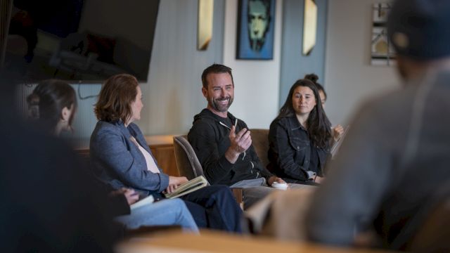 A diverse group in a meeting room, people seated in a circle, one man speaking with hand gestures while others listen attentively.