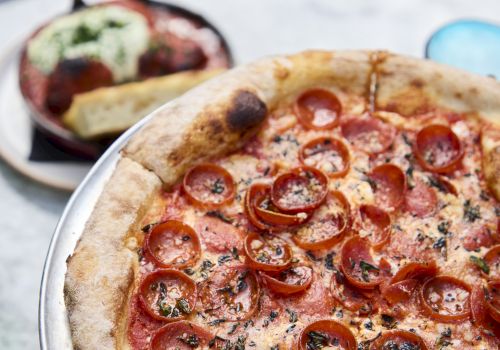 A pepperoni pizza with herbs sits on a metal tray, with a side dish and glassware in the background, ready for sharing.