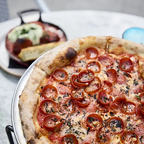 A pepperoni pizza with herbs sits on a metal tray, with a side dish and glassware in the background, ready for sharing.