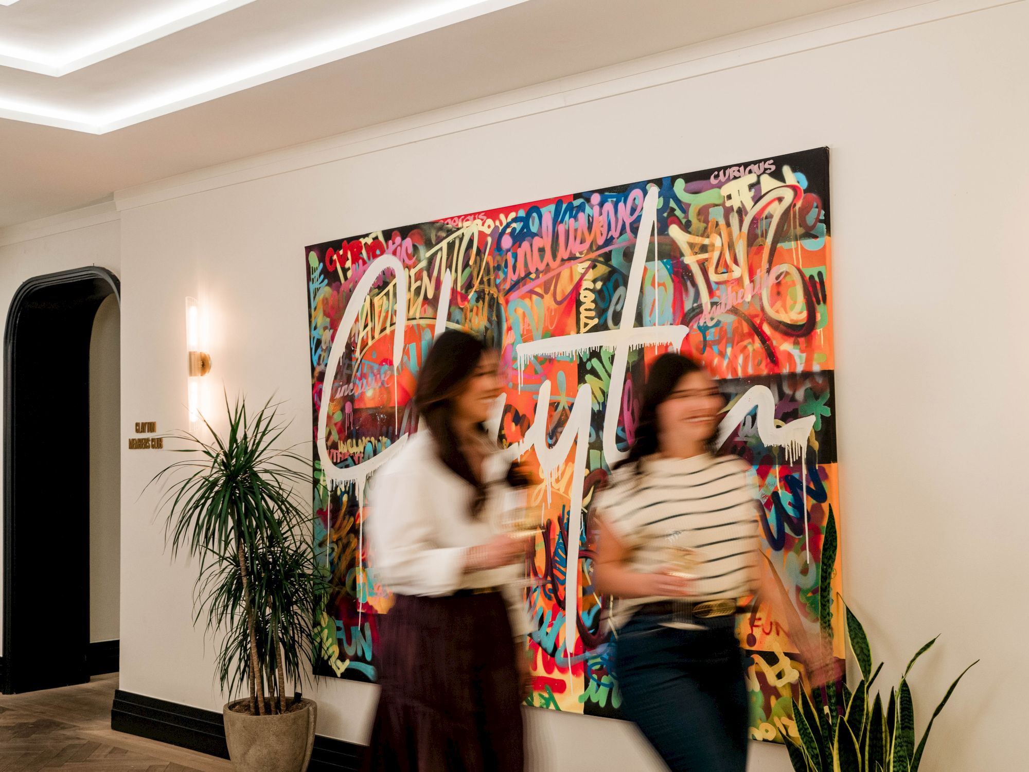 Two blurred women walk past a large colorful graffiti-style mural in a modern hallway with plants, creating a lively, urban vibe.