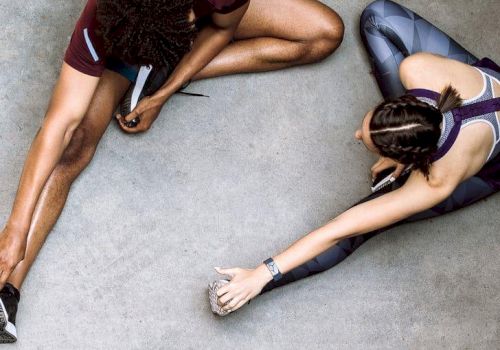 Two people rest after a workout, sitting on a concrete floor and stretching toward opposite sides, legs extended and ankles close together.