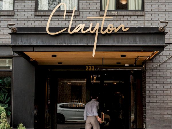 A person walks into a trendy building with a neon &ldquo;Clayton&rdquo; sign above the entrance, gray brick exterior, and a rug at the doorway.