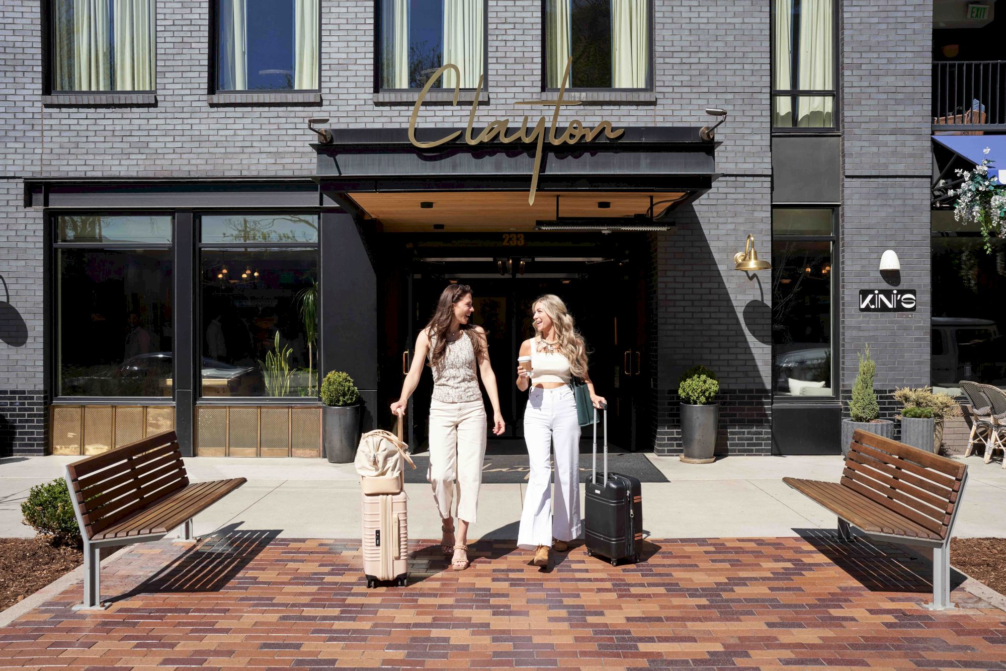 Two women walk with luggage outside a modern brick hotel, smiling and chatting on a sunny day in front of a bench-lined brick plaza.