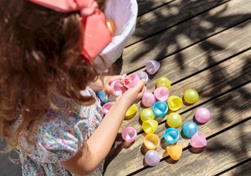 A child with a pink bow picks up colorful plastic Easter eggs scattered on a wooden deck, wearing a floral dress and short sleeves.