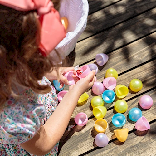 A child with a pink bow picks up colorful plastic Easter eggs scattered on a wooden deck, wearing a floral dress and short sleeves.